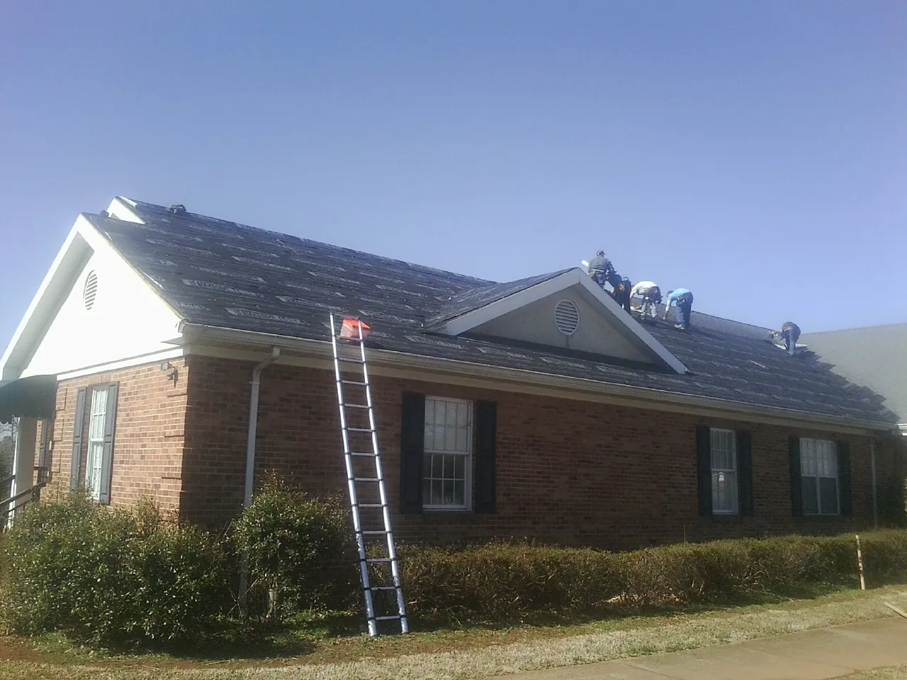 Crew tearing shingles off a brick ranch roof with a ladder set — Emergency Roof Repair work by our crew in Ellenwood