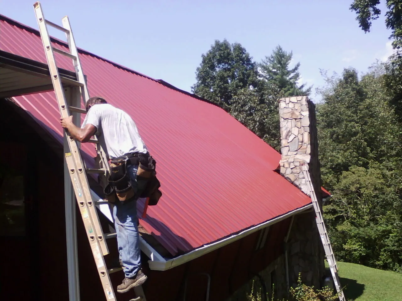 Roofer on a ladder beside a red metal roof and stone chimney — Soffit & Fascia Repair by Roofing Brothers in Ellenwood